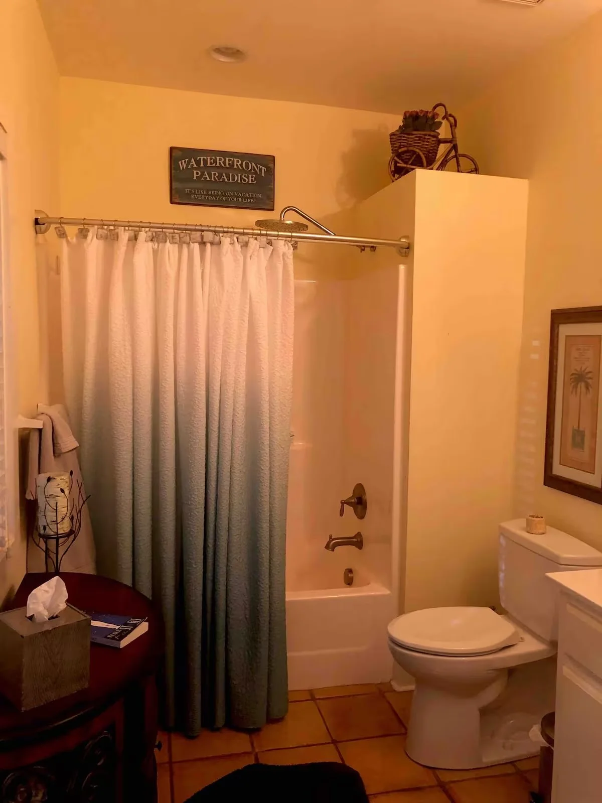 Bathroom with shower, toilet, framed artwork, towel, and decorative items on a shelf above the partition wall. Sign reading 'Waterfront Paradise' on the wall.