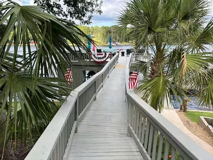 A wooden boardwalk leading to a dock with boats, decorated with American flags and bunting, surrounded by palm trees.