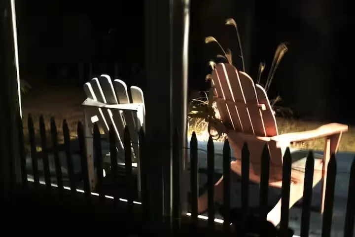 Two Adirondack chairs, one white and one brown, placed outside at night behind a black picket fence.