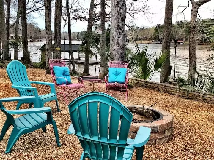 Colorful outdoor seating area with turquoise and pink chairs around a fire pit near a lake, with trees and a bridge in the background.