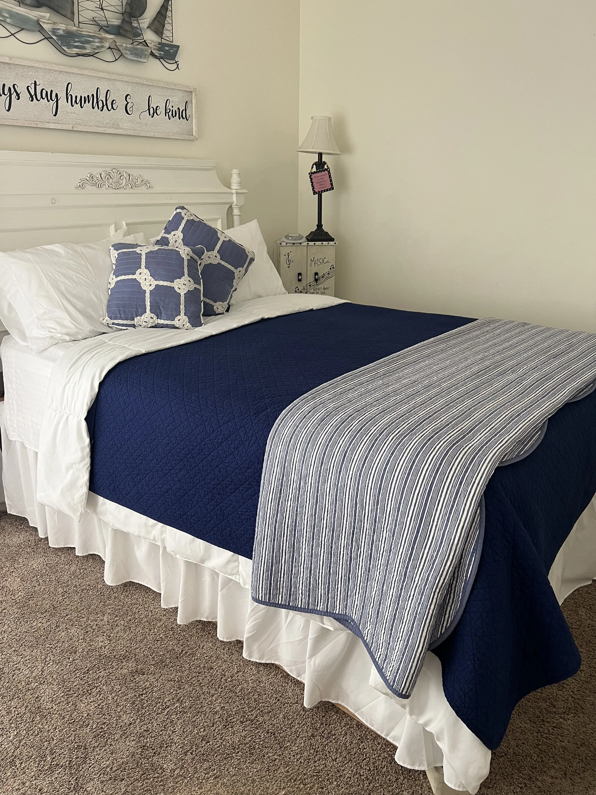 A neatly made bed with white, blue, and striped bedding. There are two matching blue pillows with white crochet design. A white bed skirt hangs down to the carpeted floor. Next to the bed is a white nightstand with decorative music-themed artwork and a small clock. A black lamp with a white shade and a pink sign hangs on the wall near the lamp. A wall sign reads, 'Always stay humble & be kind,' with a decorative nautical-themed wall hanging above it.
