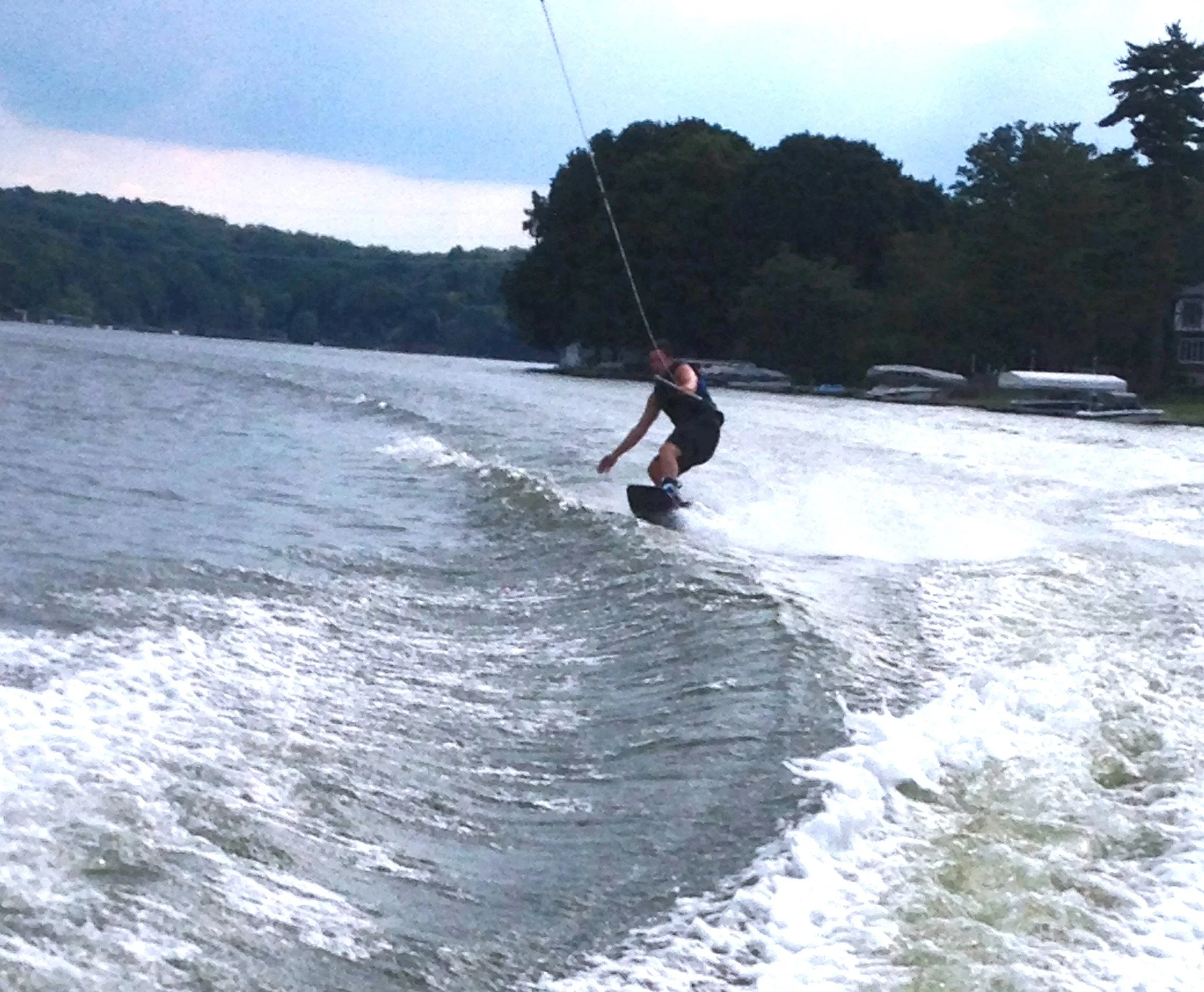 Person wakeboarding on a lake during dusk, with trees and boats in the background.