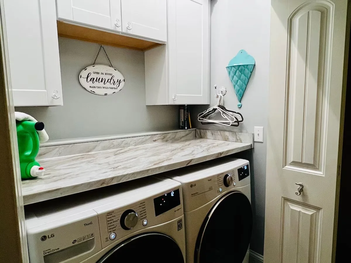 A laundry room with white cabinets, a marble countertop, a washing machine and dryer, a laundry sign, and decorative ice cream cone wall decor.
