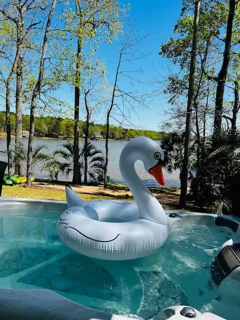 An inflatable swan float in a backyard hot tub near a lake, with trees and a clear blue sky in the background.