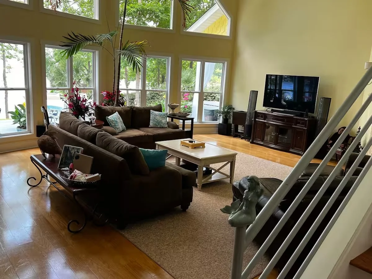 A living room with large windows, yellow walls, and hardwood floors. It has a brown sectional sofa with pillows, a white coffee table, a TV on a wooden stand, and various plants and decorations. A staircase with a decorative lion figure is partially visible in the foreground.