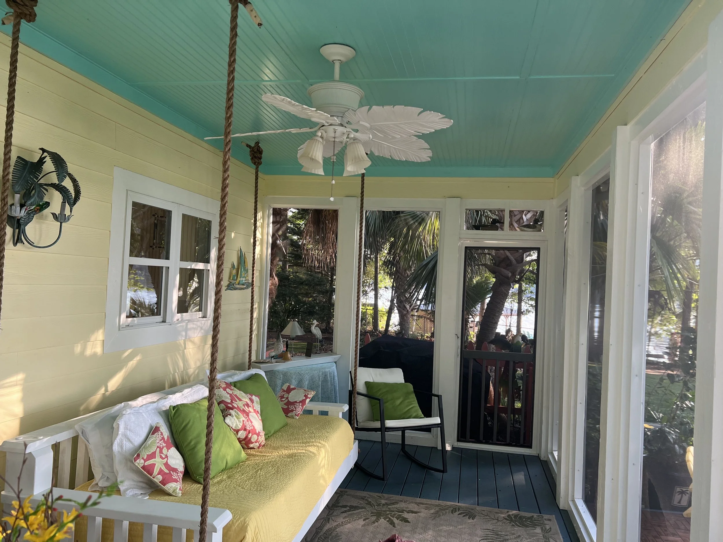 Sunroom with yellow walls, teal ceiling, ceiling fan with leaf-shaped blades, swing with green and pink pillows, and large windows showing outdoor trees.