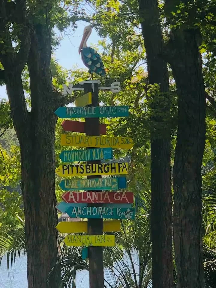 Colorful signs with city and state names, including Maumee Ohio, Stuart Florida, Pittsburgh Pennsylvania, Atlanta Georgia, Anchorage Alaska, Manhattan, Lake Mohawk New Jersey, Honolulu Hawaii, and others, mounted on a pole among trees near a body of 