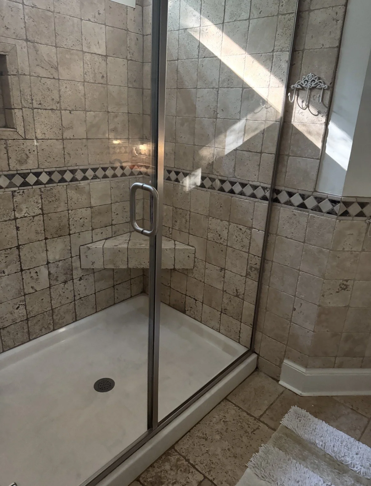 A tiled shower with glass doors, a built-in corner seat, and decorative border tiles. There is a small white rug outside the shower.