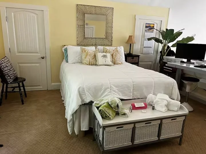 A cozy bedroom with a neatly made white bed, decorative pillows, a mirror above the bed, a nightstand with a lamp, a tall plant, a TV on a small desk, and a storage bench at the foot of the bed with towels and toiletries on top.