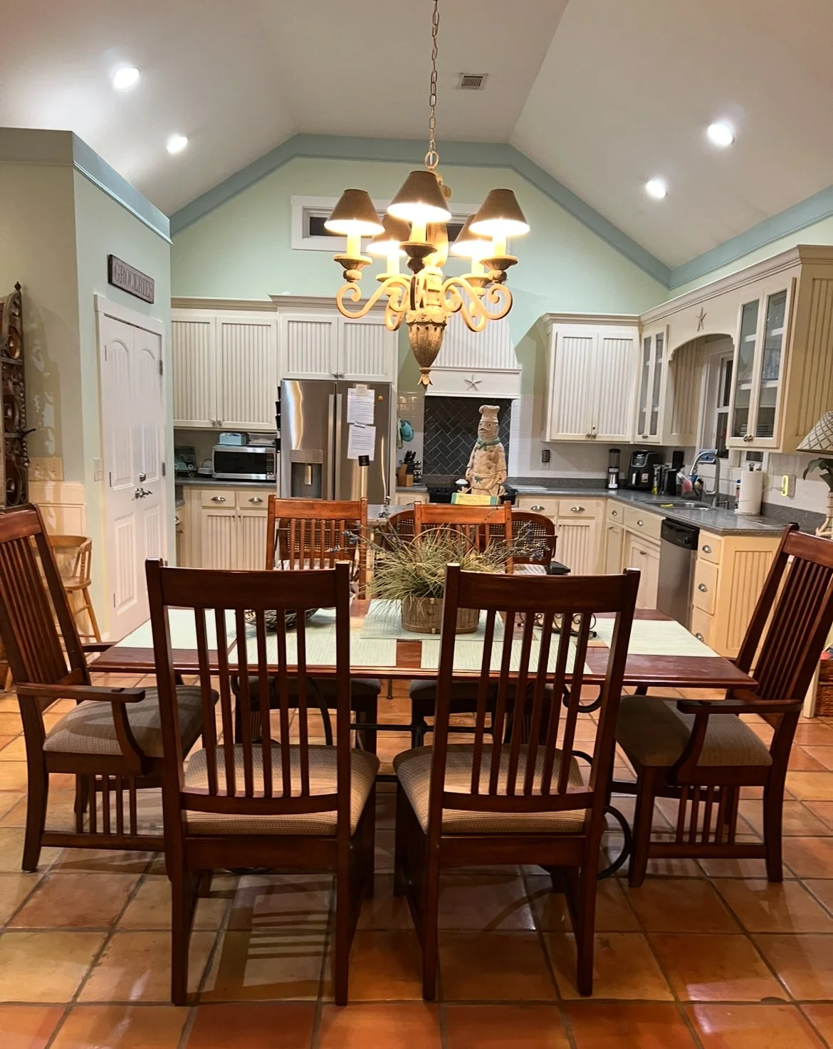 Kitchen with white cabinets, stainless steel refrigerator, black counter, dining table with six wooden chairs, chandelier overhead, decorative plant centerpiece, and various kitchen appliances.