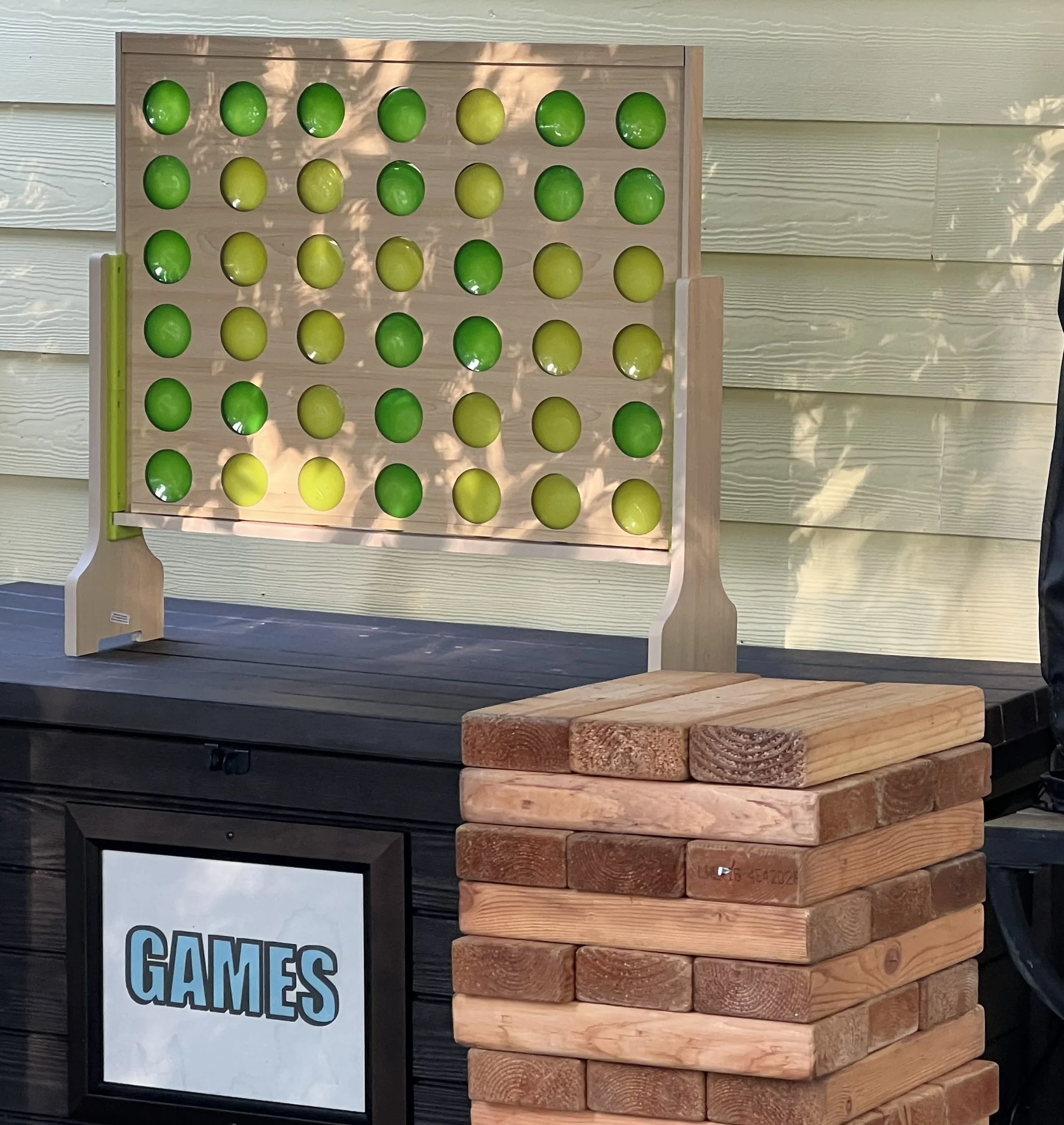 Stacked wooden blocks with some blocks supporting a wooden game board with green and yellow game pieces, and a sign with the word 'GAMES' in the background.
