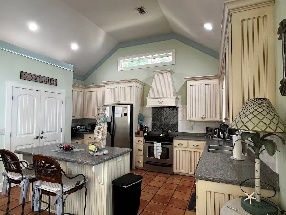 A kitchen with cream-colored cabinets, a black oven and stove, stainless steel refrigerator, and granite countertops. There are three chairs at a small island, a Groceries sign on the wall, and various decorative items, including a lamp with a seashell design.