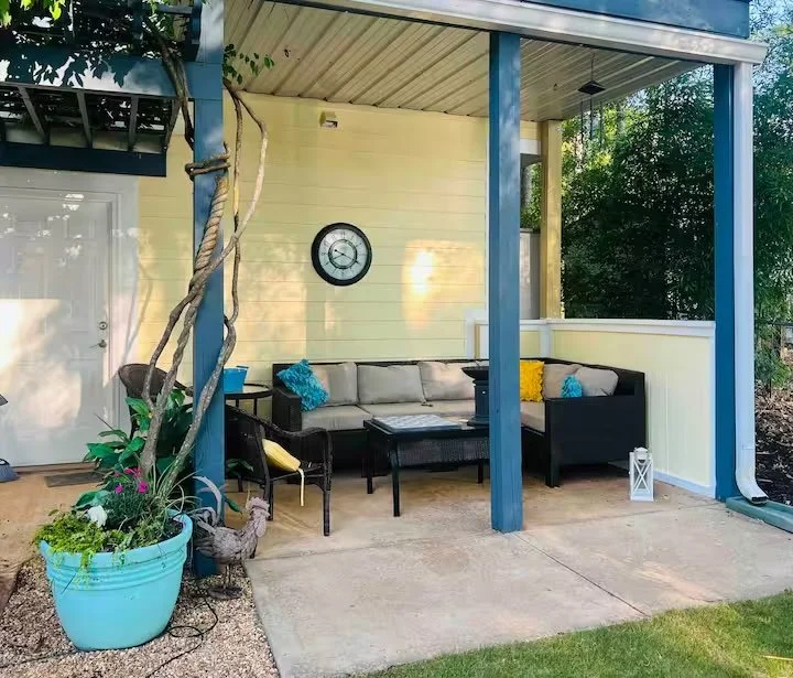 Outdoor patio with a cushioned sectional sofa, a coffee table, decorative cushions, and a potted plant on the left side. A wall clock is mounted on the yellow wall. There is a lantern on the right side near the railing, and the area is shaded by a roof with trees in the background.
