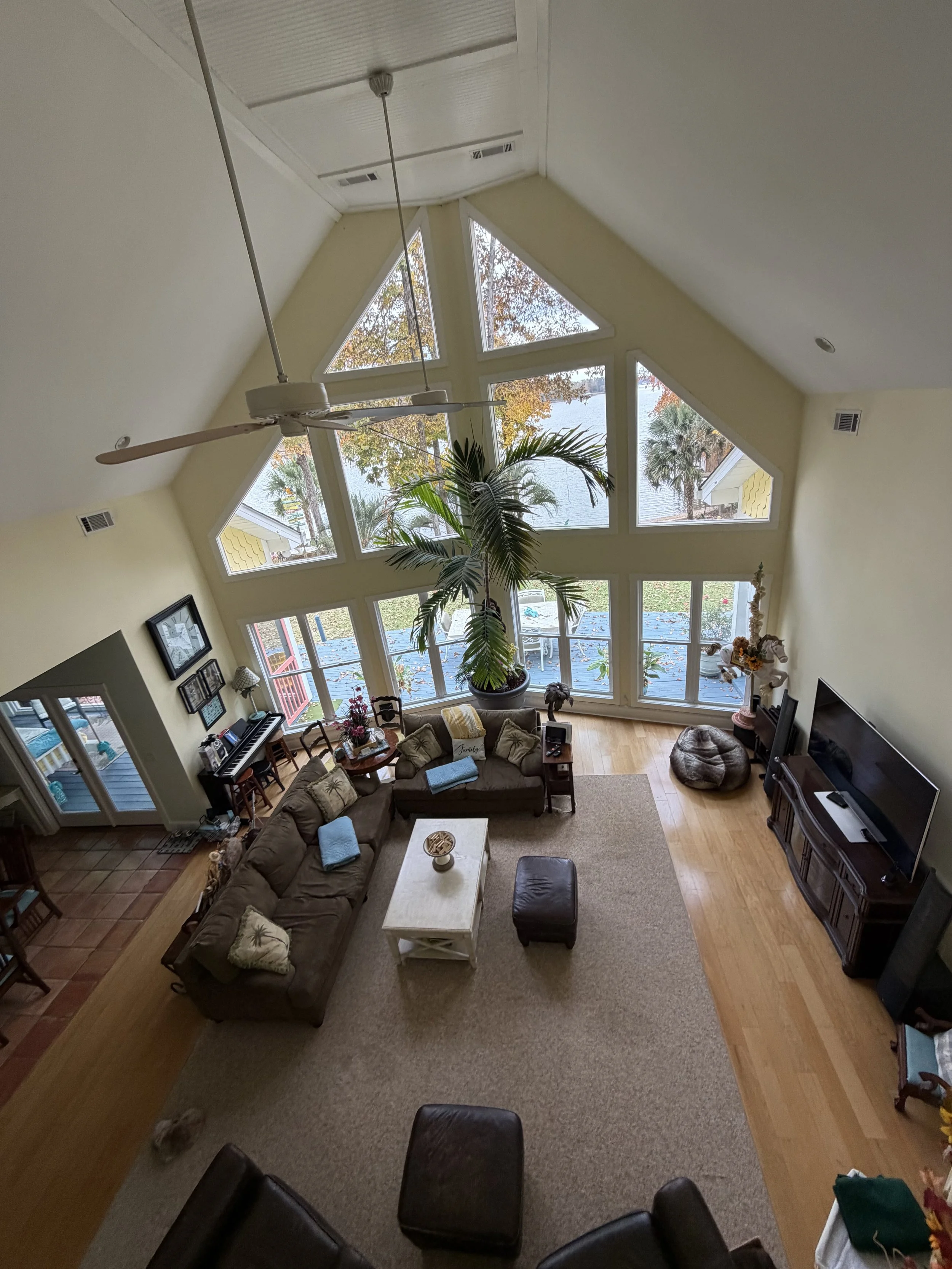 Living room with high, angled ceiling featuring large triangular windows, a ceiling fan, a sectional sofa with throw pillows, a coffee table, a large potted plant, and a view of the outdoors with trees and water.