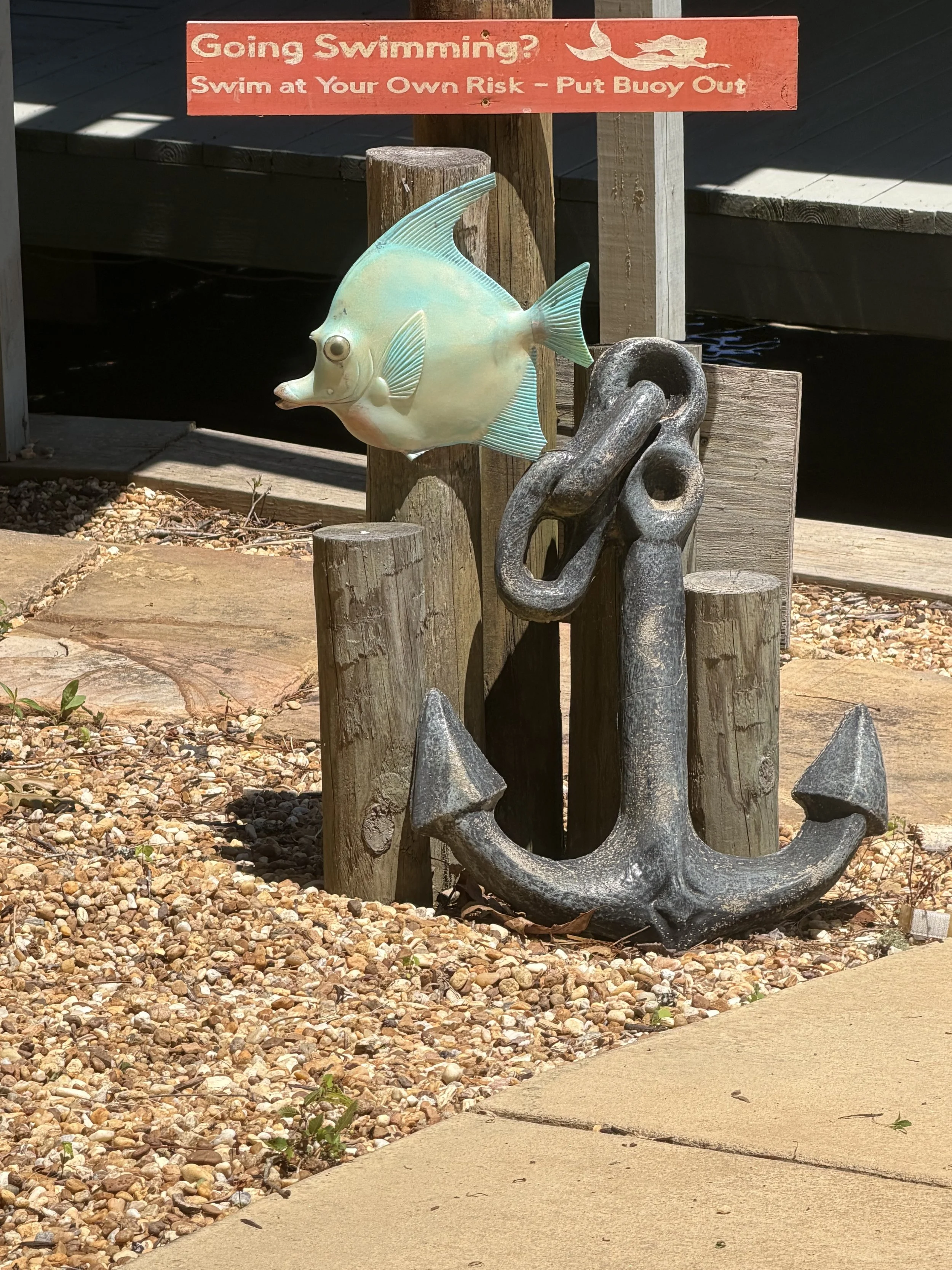 A decorative metal fish sculpture attached to wooden posts next to a large anchor sculpture, with a red sign above that reads, 'Going Swimming? Swim at Your Own Risk – Put Buoy Out'.
