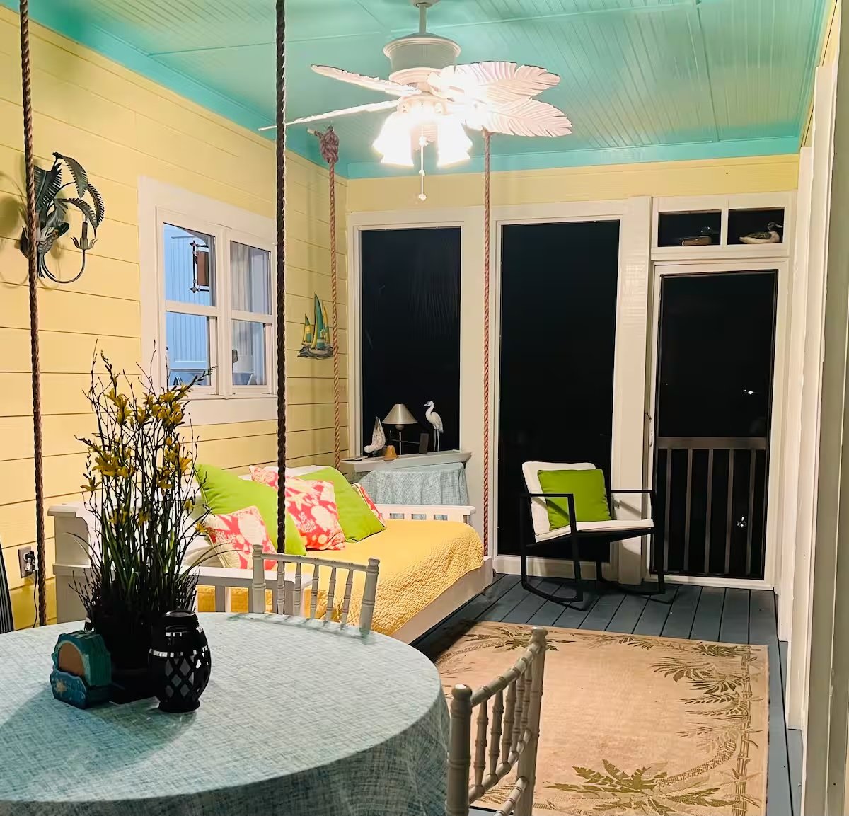 Covered porch with yellow and turquoise wall paneling, hanging swing with cushions, round dining table with flowers and decor, sitting area with chairs, ceiling fan with leaf design, dark screened door, and decorative nautical accents.