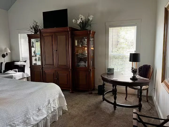 A bedroom with a bed on the left, a wooden display cabinet with glass doors in the center, a window with blinds behind the cabinet, and a round wooden table with a lamp on it to the right. There's a side chair and a TV on top of the cabinet.