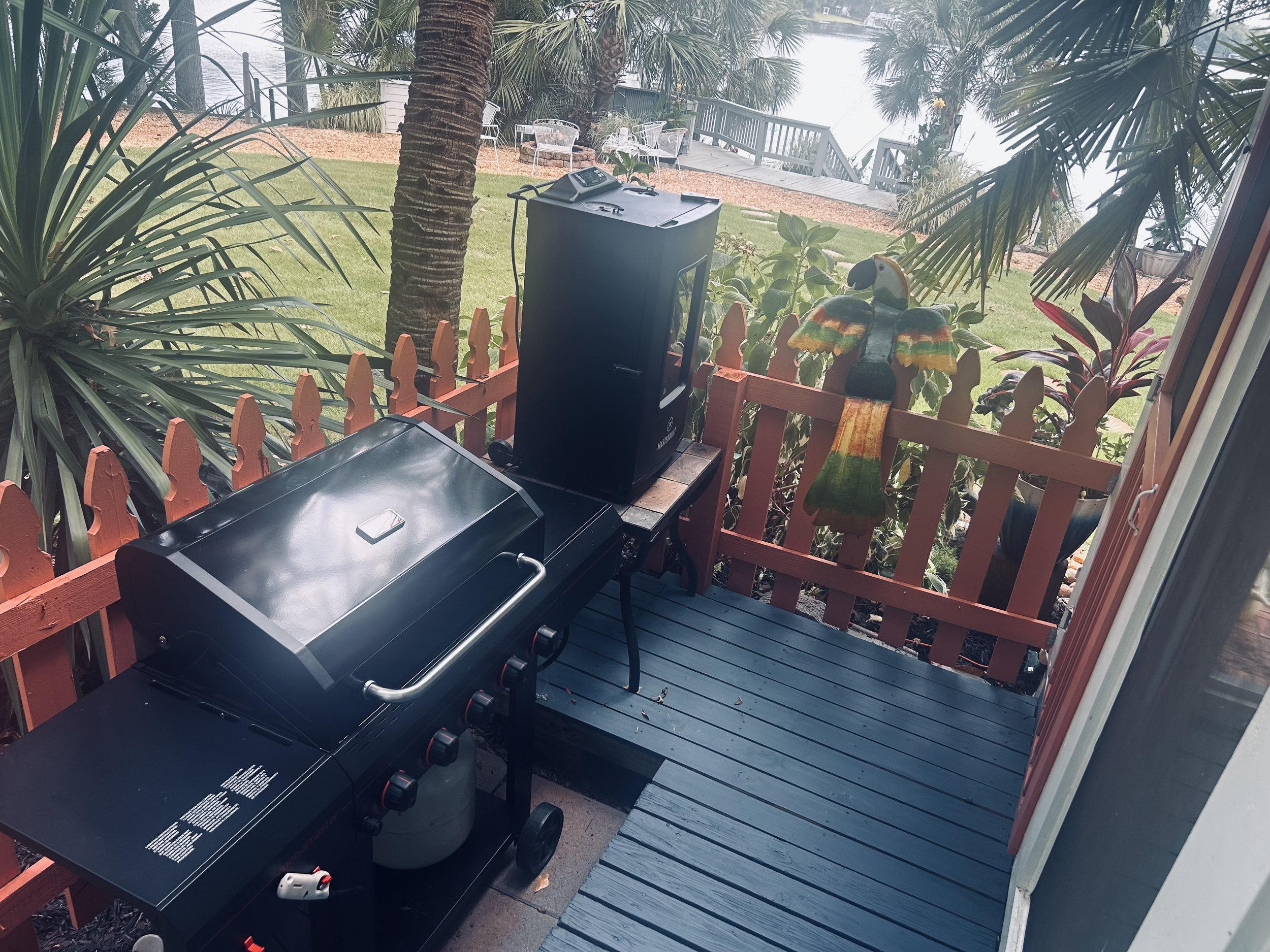 View of a backyard through a doorway, featuring a small black BBQ grill on a wooden deck, surrounded by tropical plants and trees, with a grassy yard, outdoor seating, and a raised wooden deck in the background.