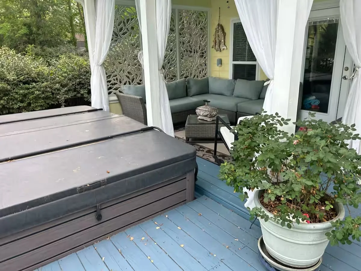 A covered outdoor patio with a sectional sofa, a wicker chair, a small side table, potted plant, and a hot tub, surrounded by greenery and white curtains.