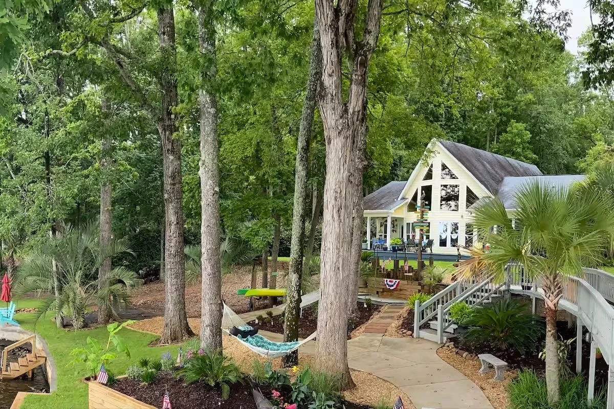 A spacious backyard with tall trees, lush greenery, a hammock, a colorful children's Adirondack chair, and a large white house with an A-frame roof and expansive windows.
