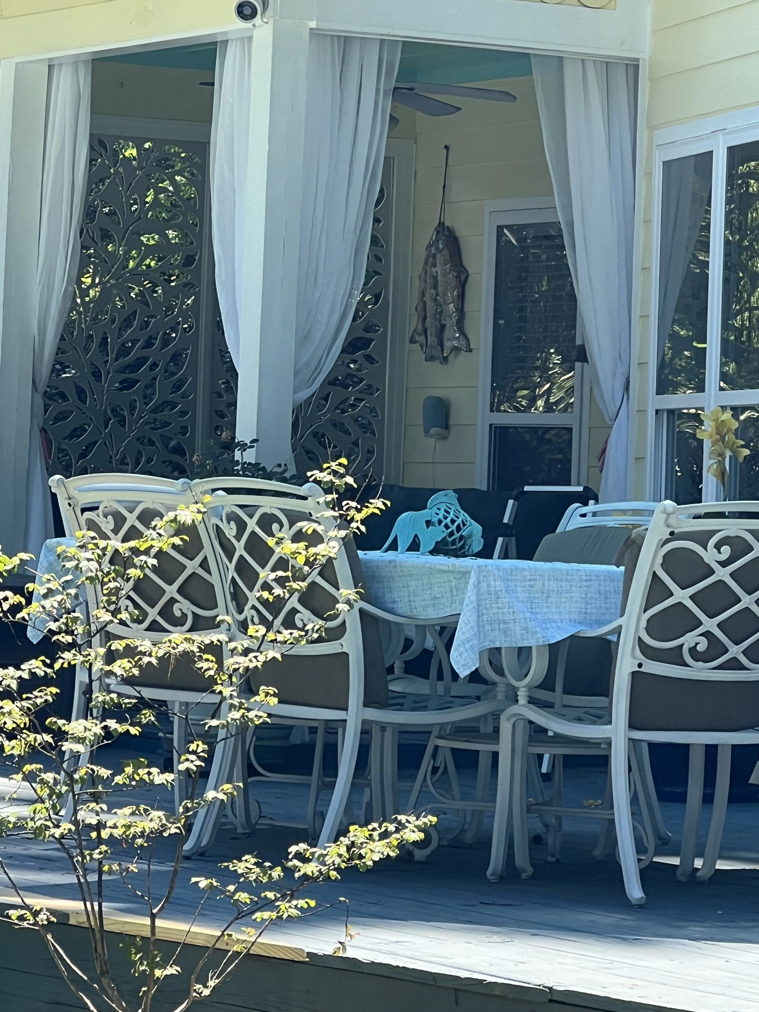 A covered outdoor patio area featuring white lattice chairs around a table with a tablecloth, a decorative wall with a fish mounted on it, white curtains, and a ceiling fan, with part of the house's yellow siding visible.