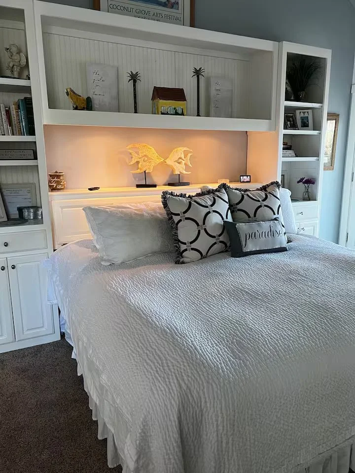 A neatly made bed with white bedding and decorative pillows, including one with the word 'paradise,' in a bedroom with built-in white shelves and a lit decorative display of two dragon sculptures.