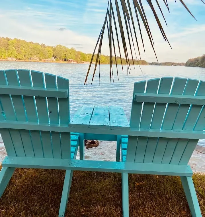 Two blue Adirondack chairs facing a lake, with a palm tree branch hanging above and a sandy beach underneath.