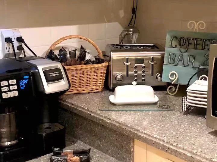 Kitchen countertop with coffee machine, bread toaster, toaster in a basket, glass jar, colorful 'Coffee Bar' sign, and a ceramic butter dish.
