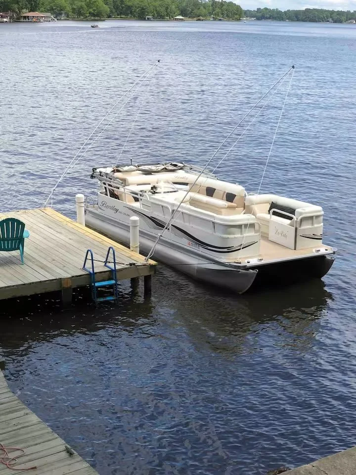A white pontoon boat docked at a wooden pier on a calm lake with trees in the background.