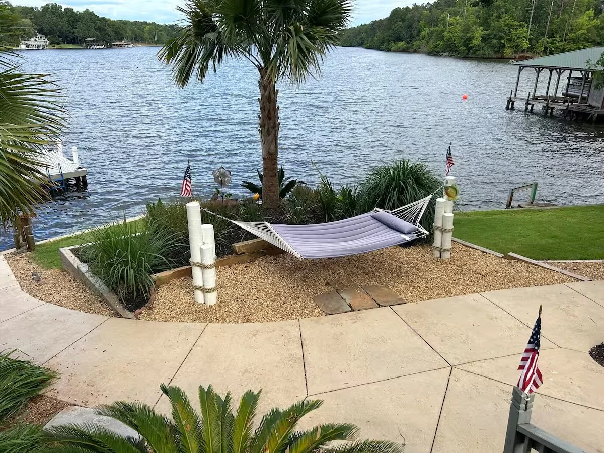 A backyard with a hammock tied between two posts, facing a river with boats and docks, surrounded by palm trees and American flags.