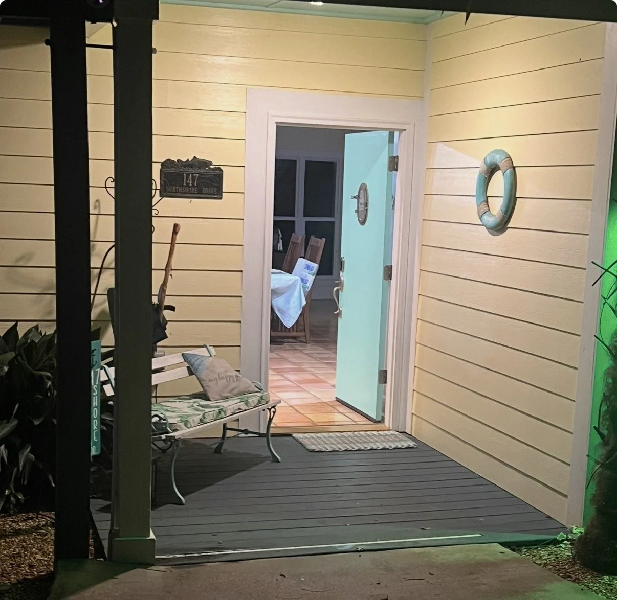 View of a house porch at night with a yellow wall, a door open revealing a dining area, a life ring hanging on the wall, a bench with cushions, and a sign on the wall displaying the house number 147.