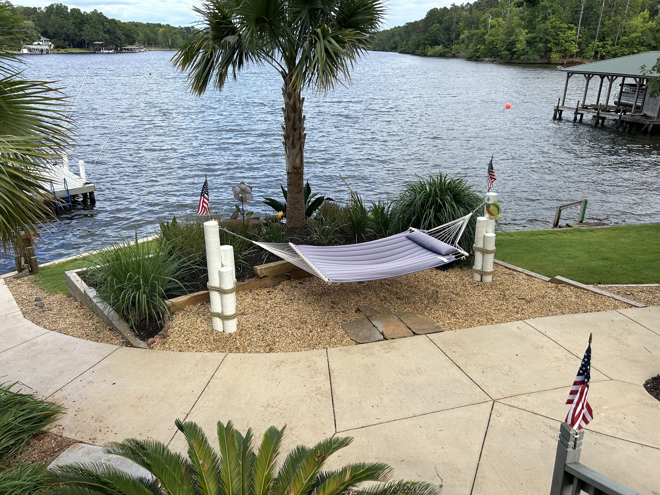 View of a lakeside with a tree, a hammock, American flags, and a dock extending into the water, surrounded by green trees.