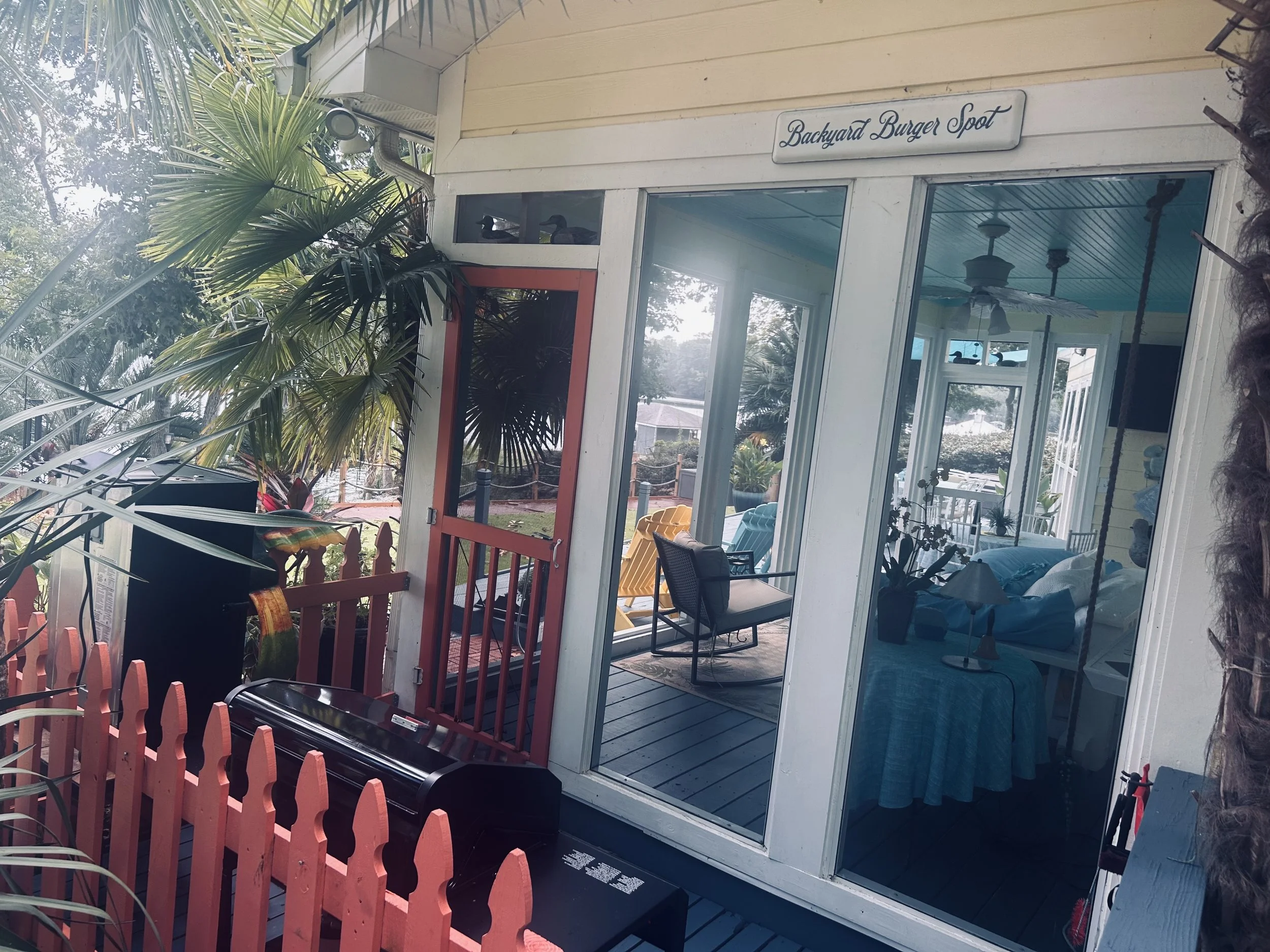 Exterior view of a building with a sign that reads 'Backyard Burger Spot,' showing large glass windows, with ducks visible inside, and a door leading to a porch area with chairs and plants. There are trees and a sky in the background.