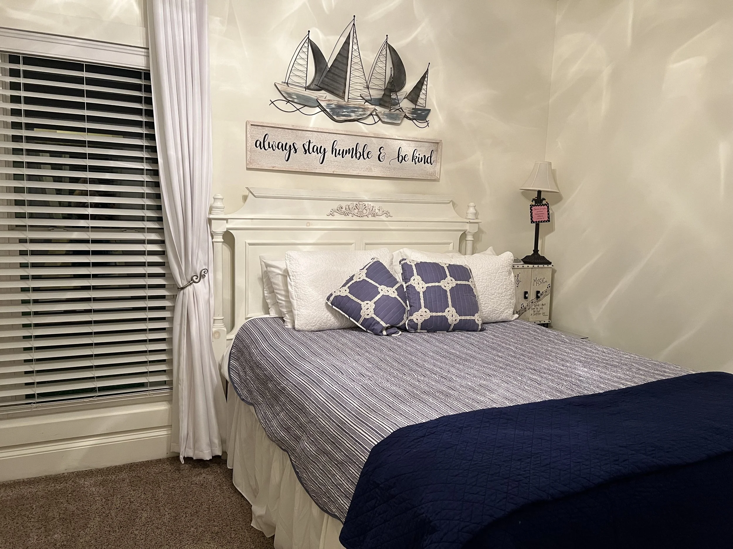 A bedroom with a white bed, decorative pillows, a nightstand, a table lamp, a window with blinds and curtains, a wall sign that reads 'always stay humble & be kind', and a metal wall art of a sailboat.