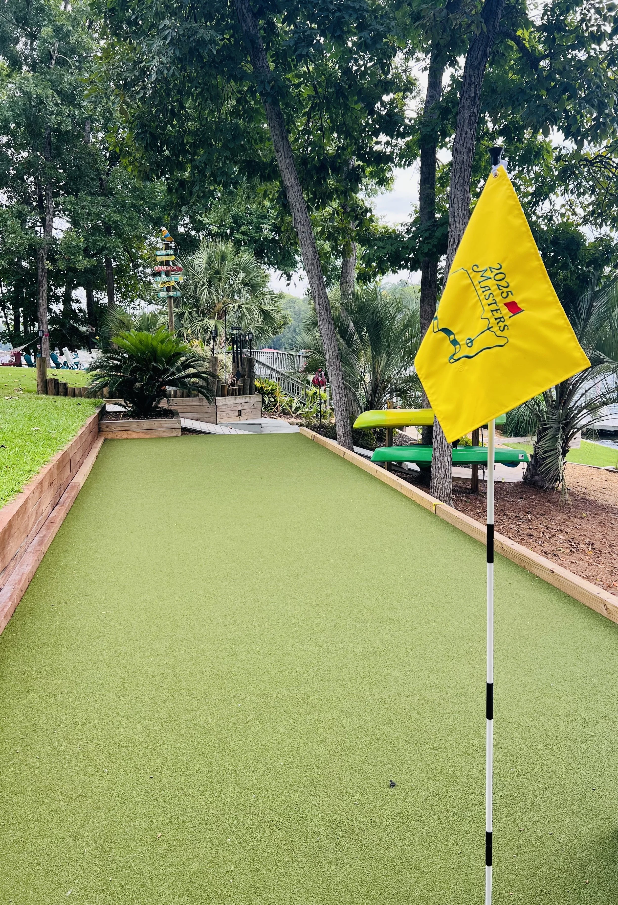 A mini golf course with a green putting surface, a yellow flag with the logo and text '2023 Masters,' surrounded by trees and plants.