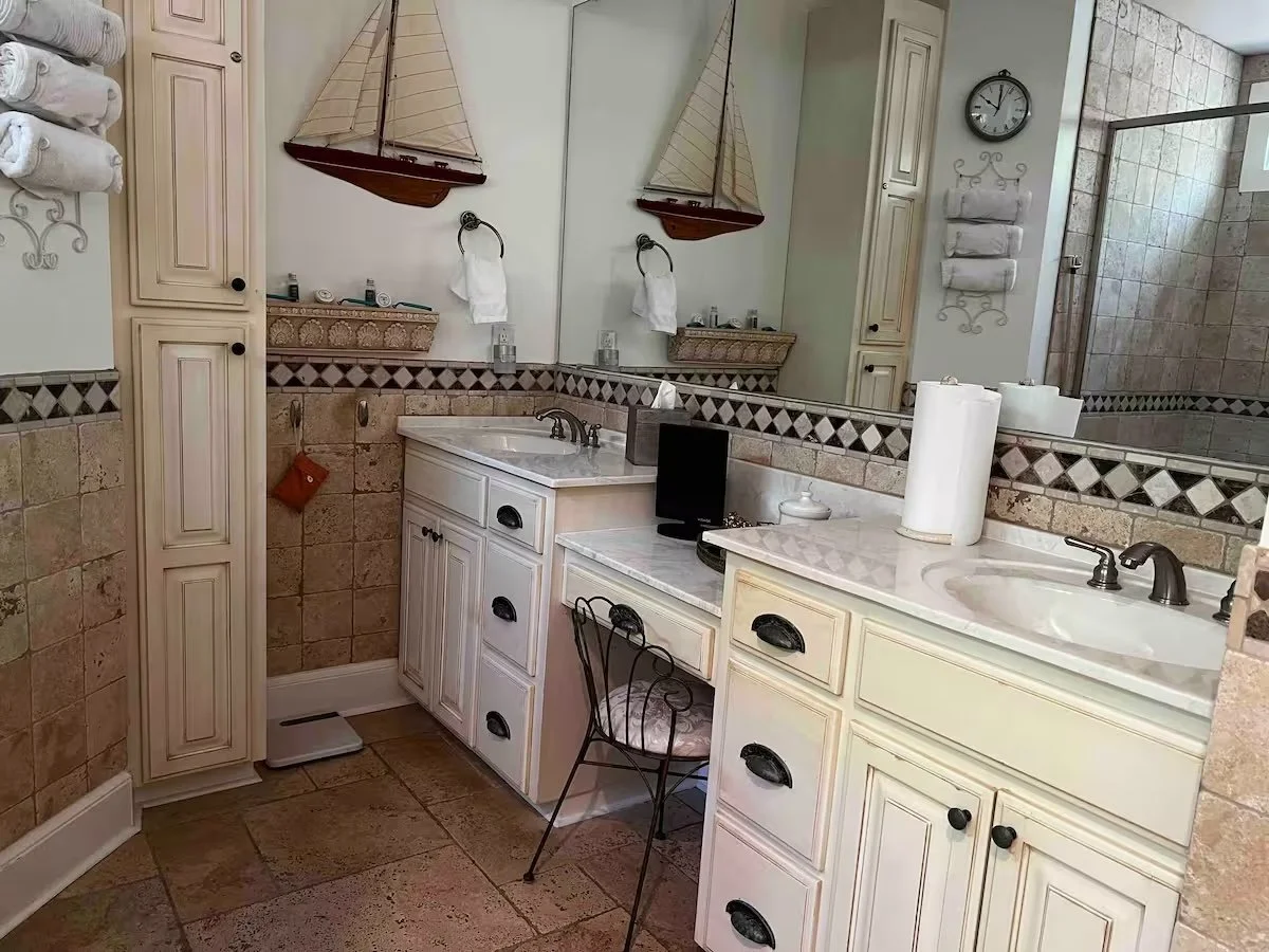 A bathroom with two sinks, large mirror, beige cabinets, nautical decorations including model sailboats, and a shower with tile walls.
