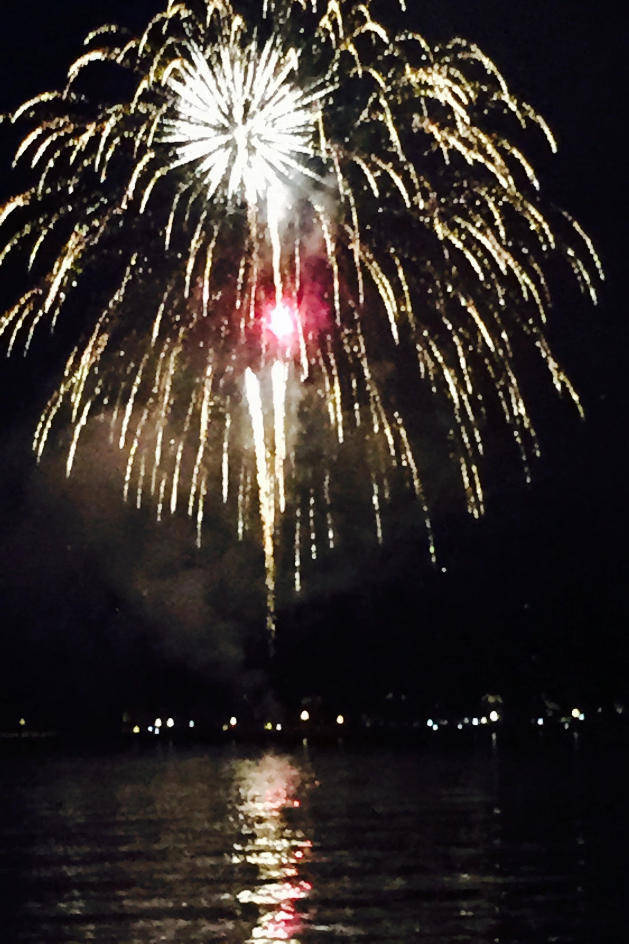 Fireworks display over a body of water at night, with reflections on the water and small lights along the shoreline.