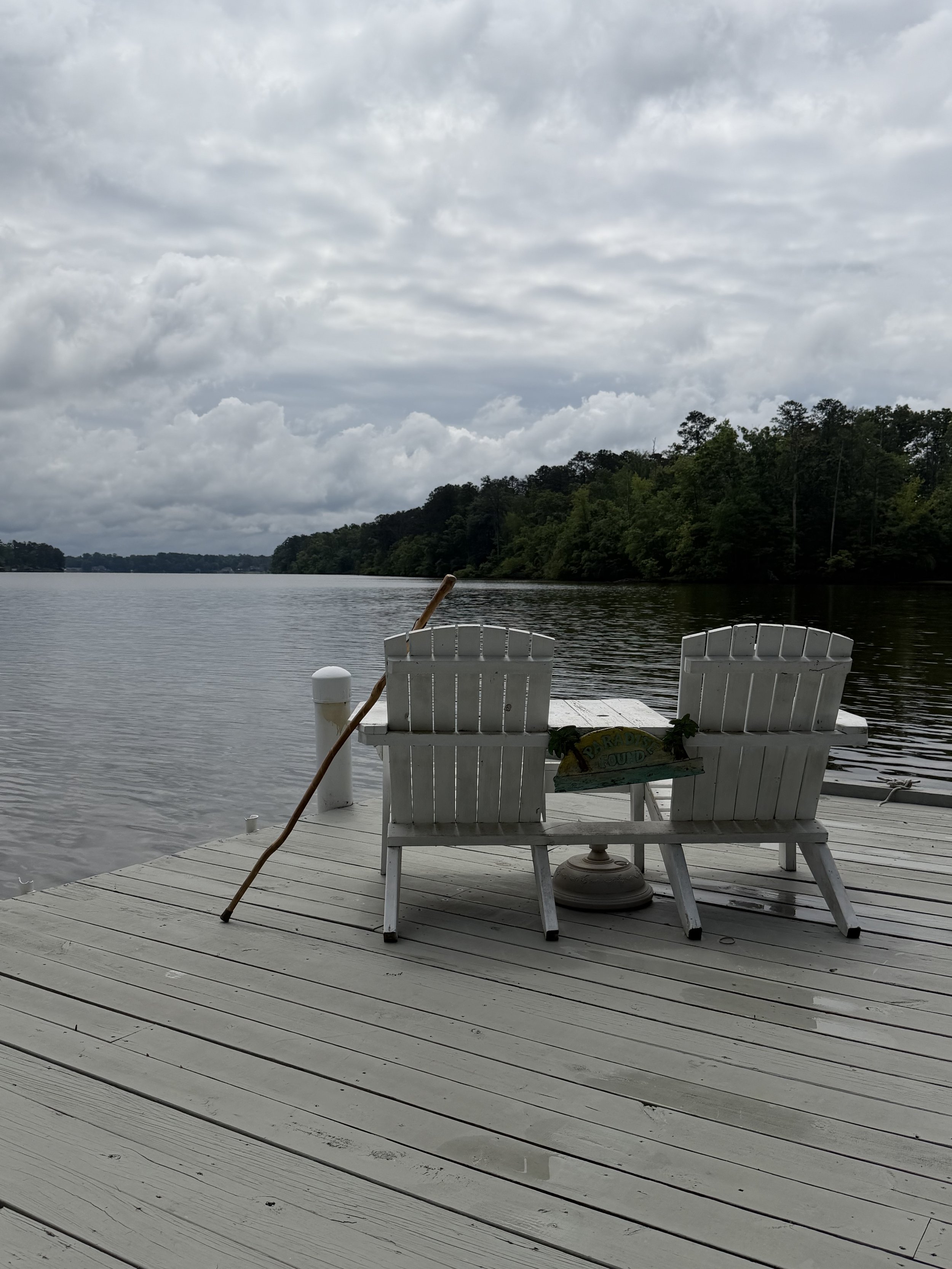 Two white Adirondack chairs on a wooden dock facing a lake with trees in the background and cloudy sky overhead.