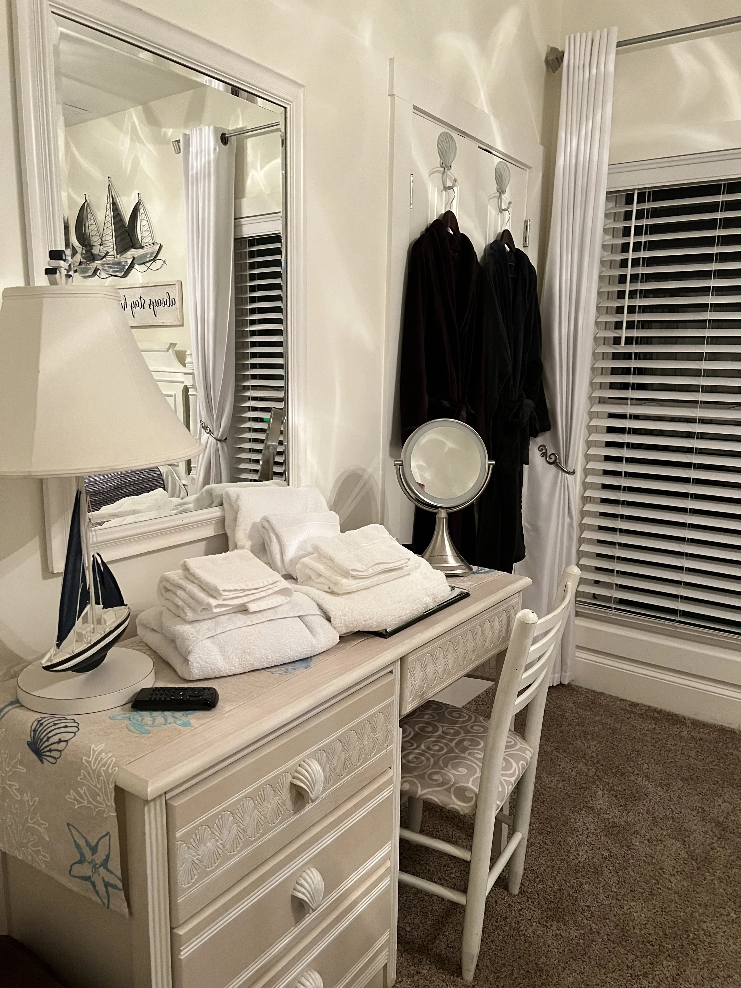 A bedroom with a dresser, a mirror, and a bed. The dresser has folded towels on top and a lamp. A mirror reflects a wall decor of sailboats. There are robes hanging on hooks near a window with blinds and white curtains.