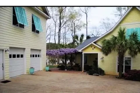 A residential driveway with two yellow houses, one with a garage door, surrounded by trees and landscaping.