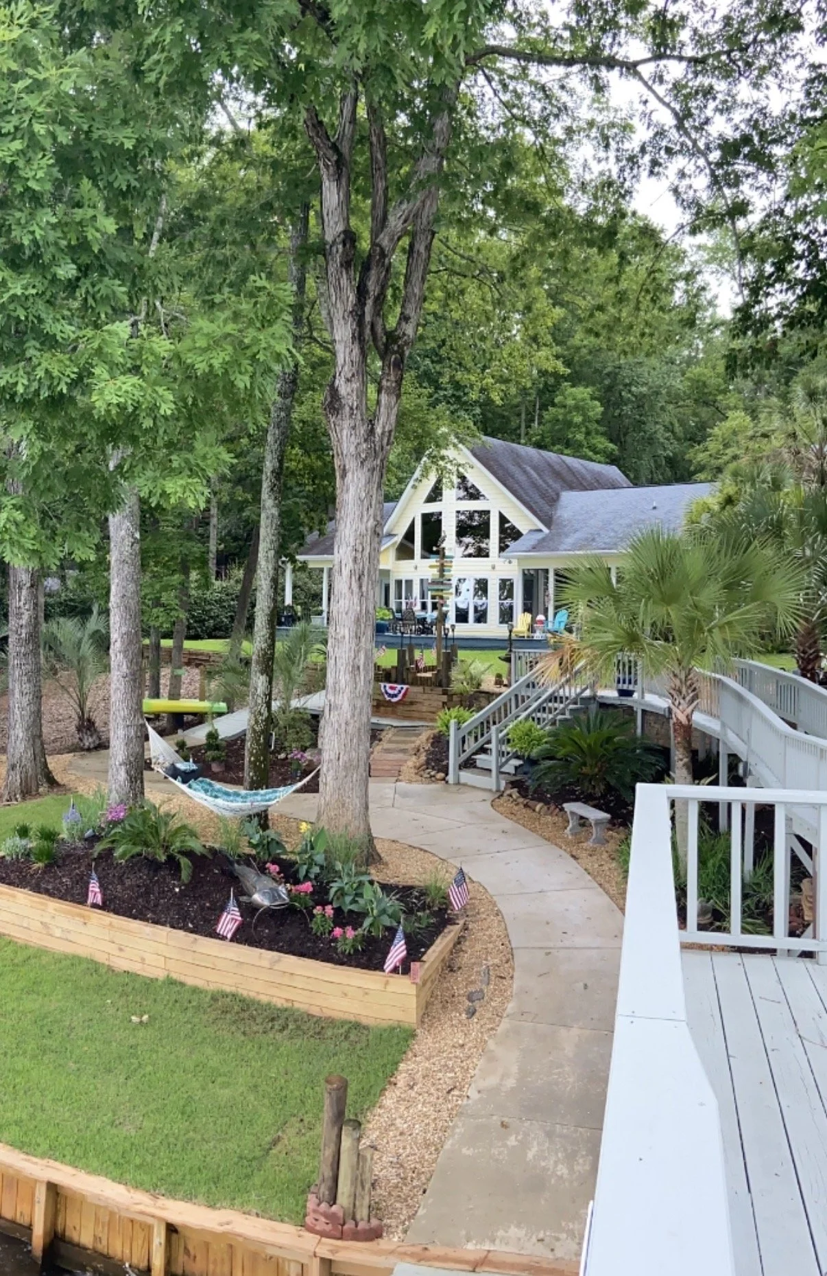 A backyard garden with a small lawn, flower beds, trees, a hammock, a walkway, and a house with large windows and a deck. Decorative flags are in the flower beds, and the area is landscaped with plants and mulch.