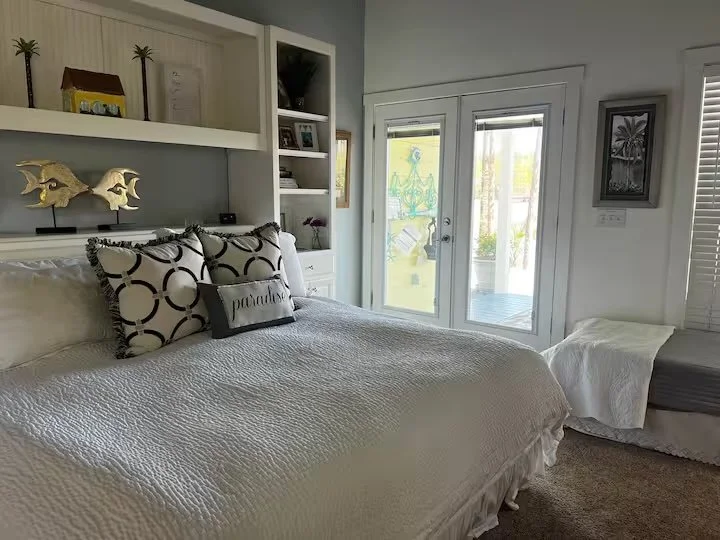 Bedroom with a large bed covered in white quilt, decorative pillows including one with the word 'paradise', built-in white bookshelf, glass door leading outside, framed picture of palm trees on wall.