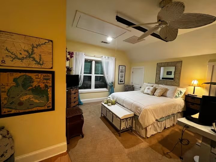 A cozy bedroom with a large bed, side table with a lamp, window with curtains, a ceiling fan with a unique leaf design, and framed pictures on the walls.