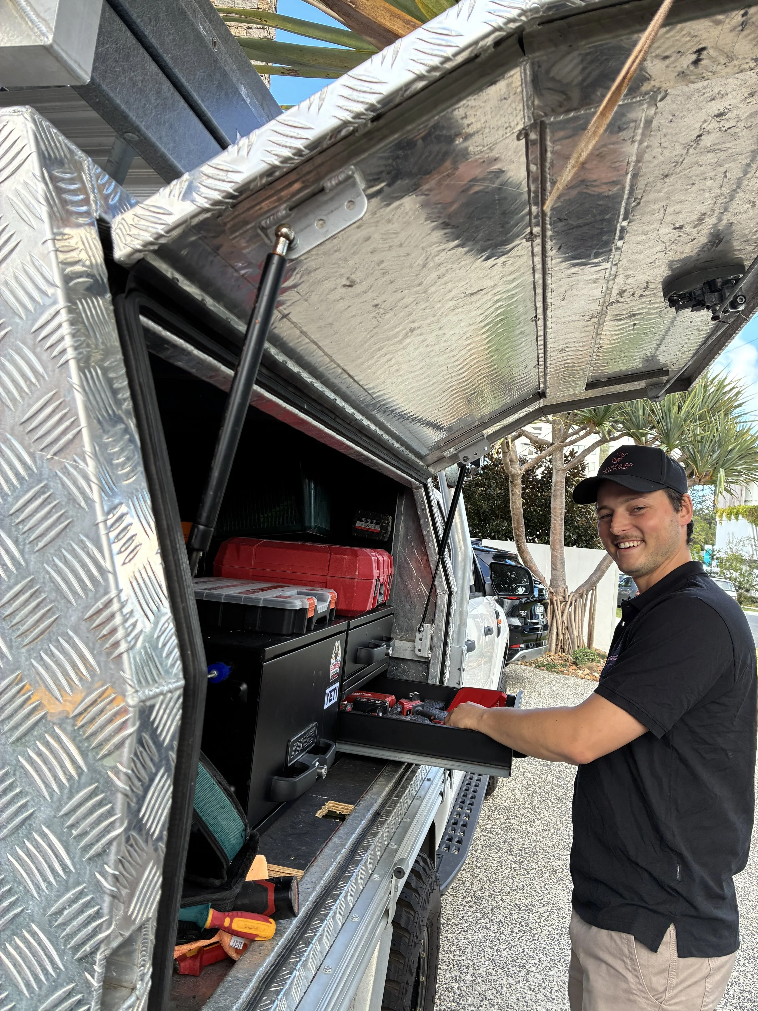 A young man smiling and wearing a black cap and black shirt is standing next to an open utility truck. Inside the truck, various tools and equipment are organized on shelves, including a red toolbox, black storage cases, and other hand tools. The truck has diamond plate metal panels and a canopy. The background shows trees and parked cars.