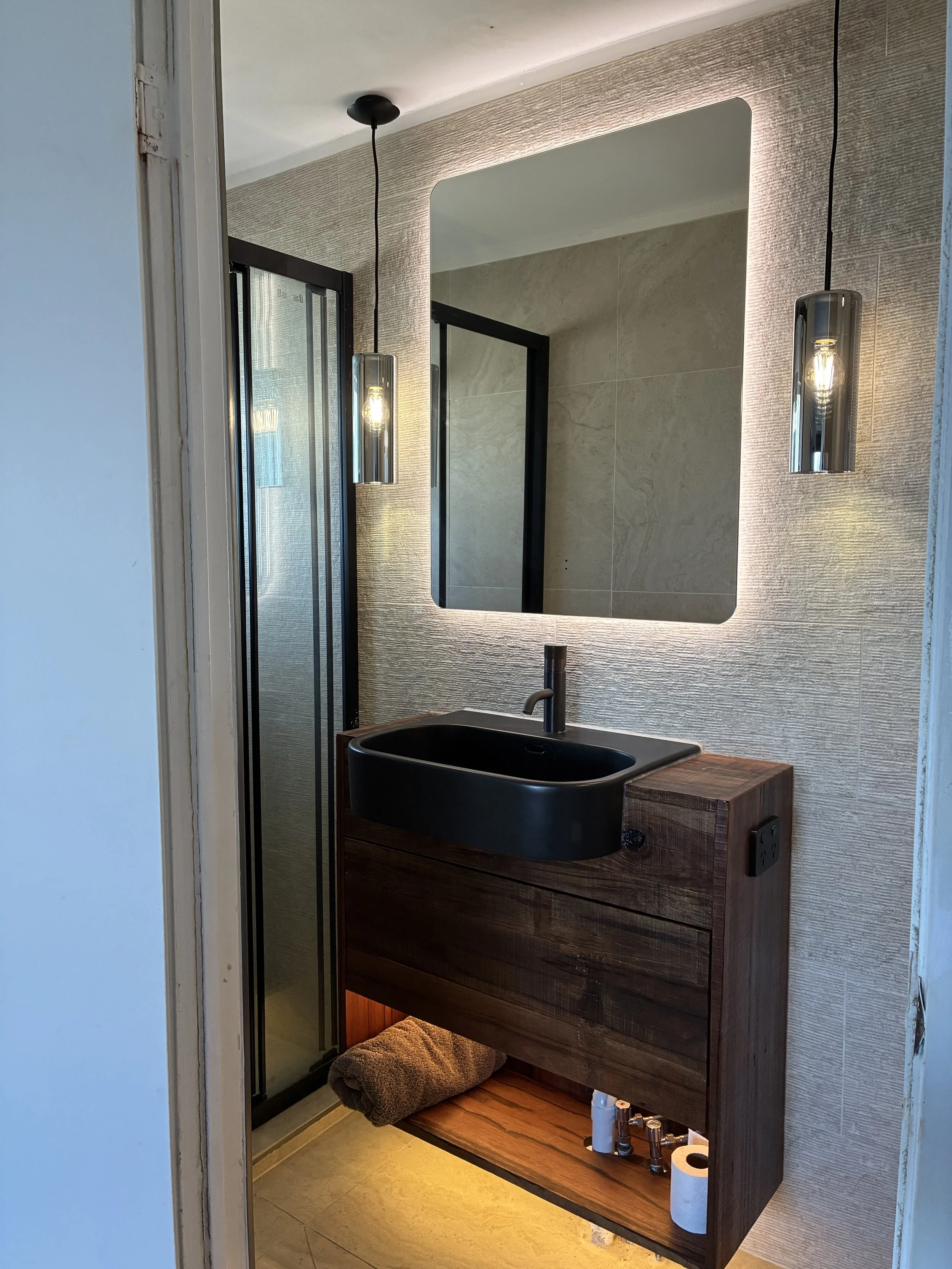 Modern bathroom with textured wall, illuminated mirror, black sink on a dark wooden vanity, hanging pendants, shower with glass door, towel on the floor, and toiletries on the lower shelf.