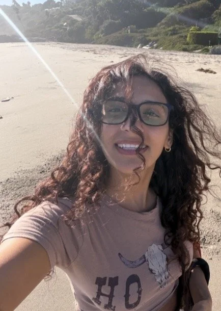 Woman with curly hair and glasses taking a selfie on a beach with the sun shining.