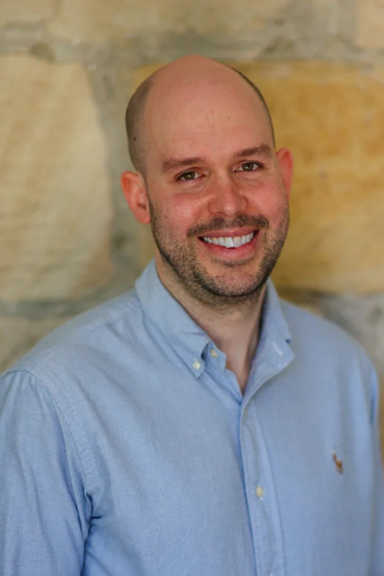 Portrait of a smiling man with a bald head, light skin, wearing a light blue button-up shirt, standing against a blurred background.