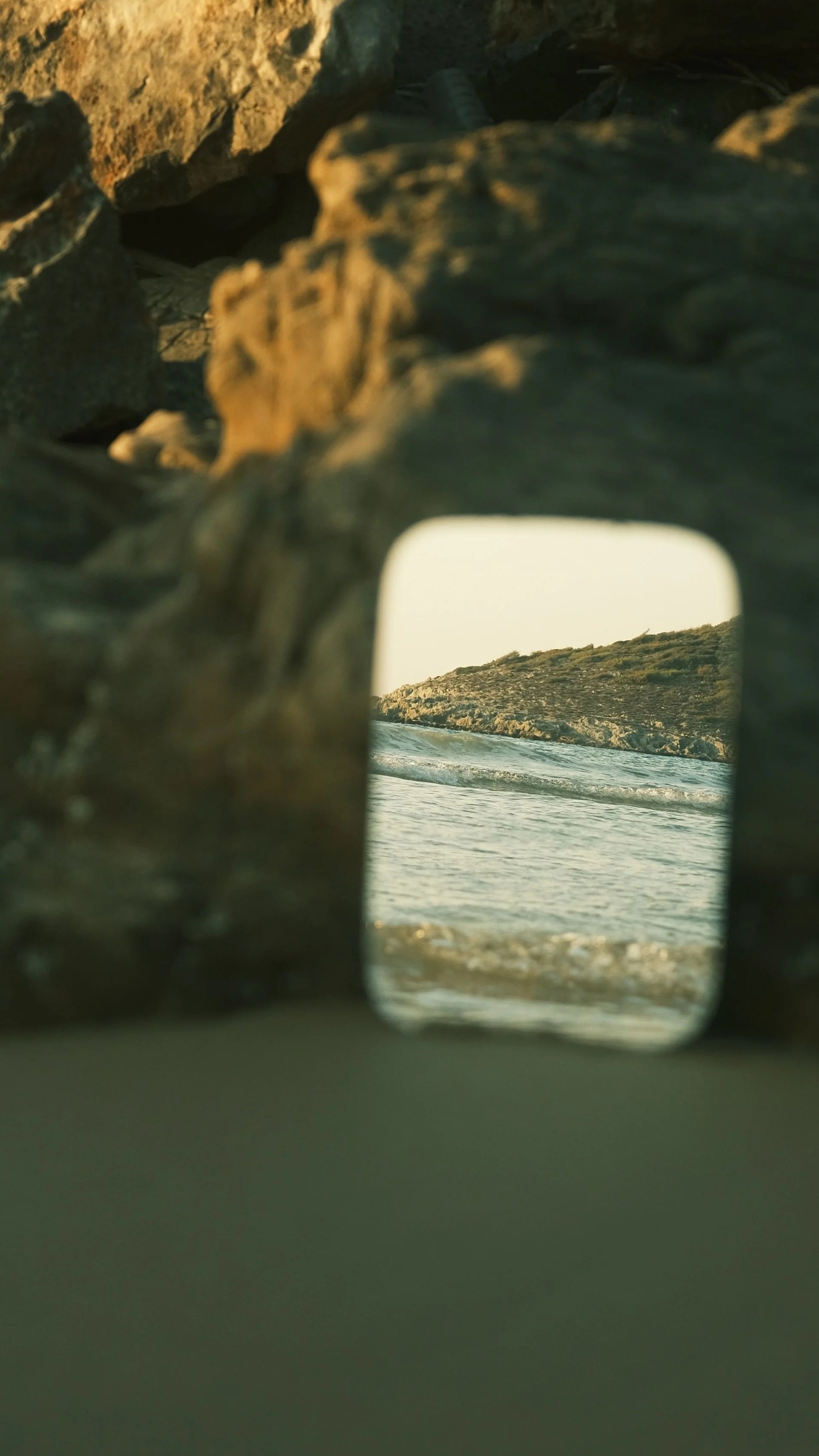 View of a coastline with waves, seen through a reflective surface in a rocky area.