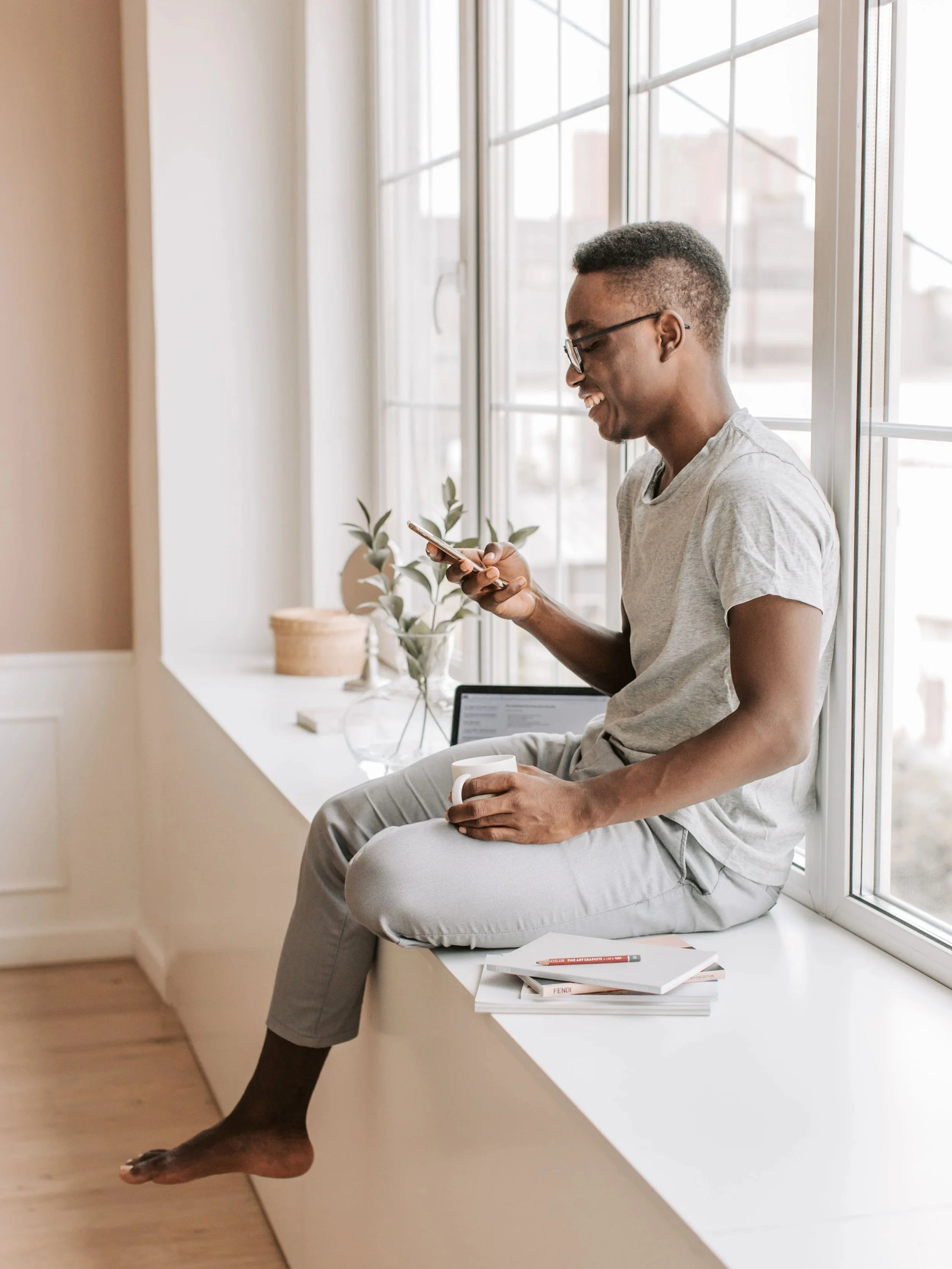 A young man with glasses, wearing a light gray t-shirt and light gray pants, sitting on a window seat, smiling while looking at his phone. He is barefoot, holding a white mug with his right hand, and has a notebook and colored pencils on a white surface beside him. The background shows large windows with a cityscape view, and a decorative plant on the windowsill.