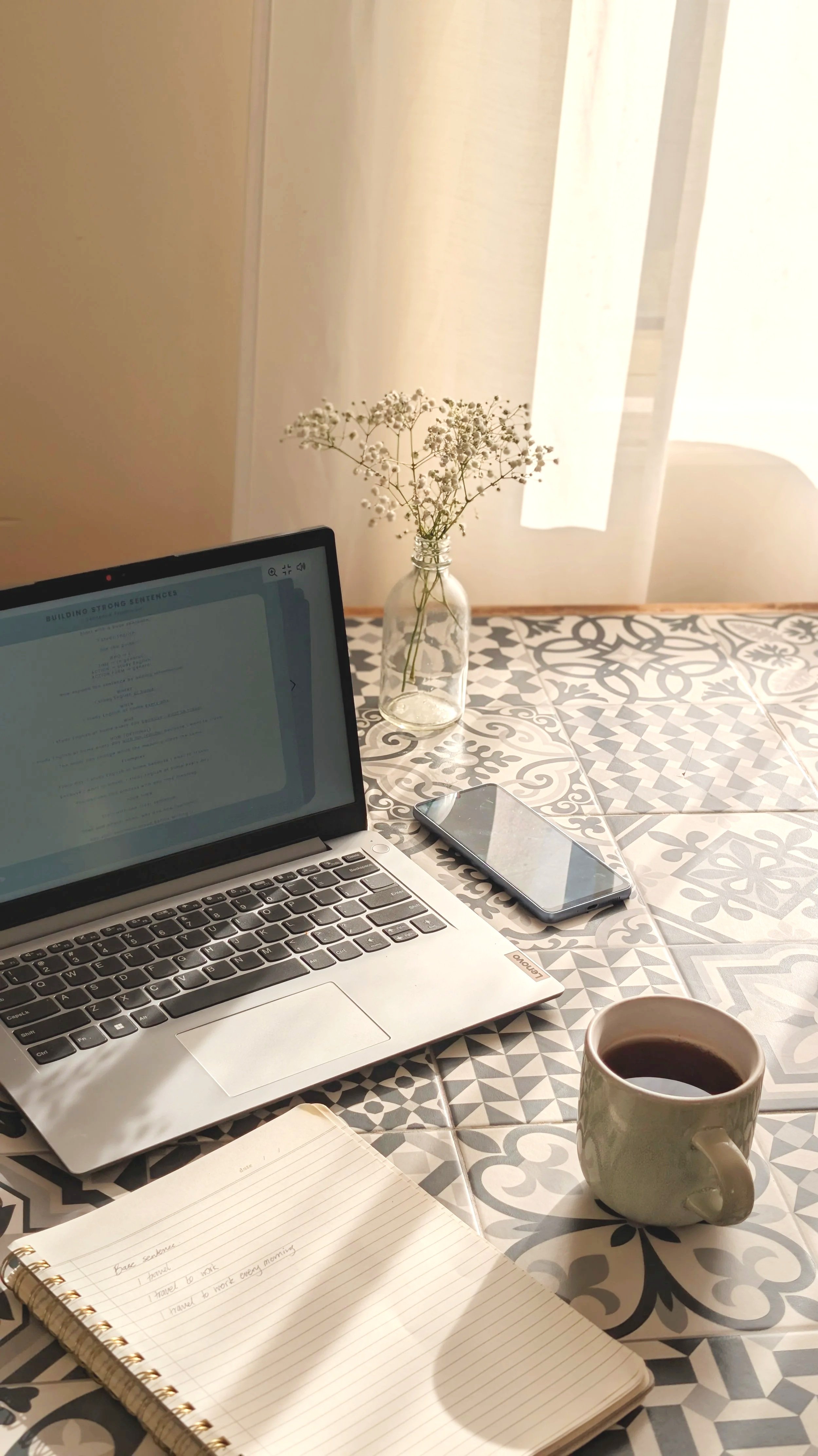 A workspace with a laptop, a smartphone, a notebook, a cup of coffee, and a vase with white flowers on a patterned table, with a sheer curtain and sunlight in the background.